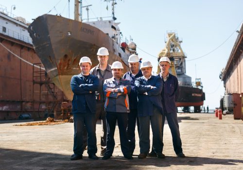Marine engineers in helmets, repair crew with vessel. Team of shipyard workers stand proudly at dry dock. Dockyard laborers by ship hull, maritime industry, maintenance on sunshine day.